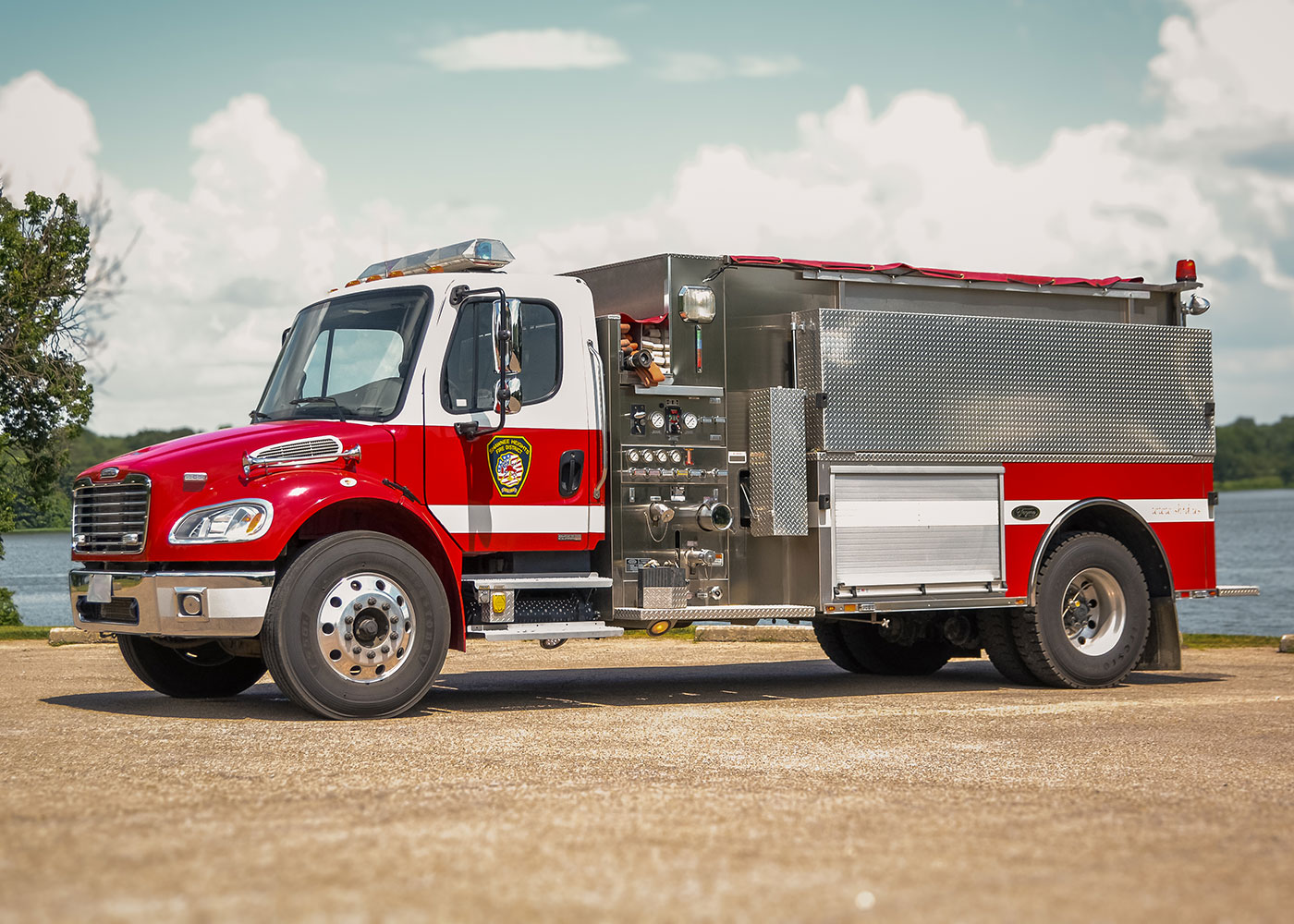A red and white fire truck with chrome details is parked on a paved surface near a body of water, with trees and a partly cloudy sky in the background.