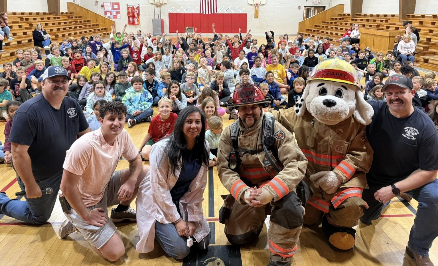 A group of firefighters, a woman in a white coat, and a person in a dog mascot costume pose with a large group of children seated on bleachers in a gymnasium. An American flag hangs in the background.