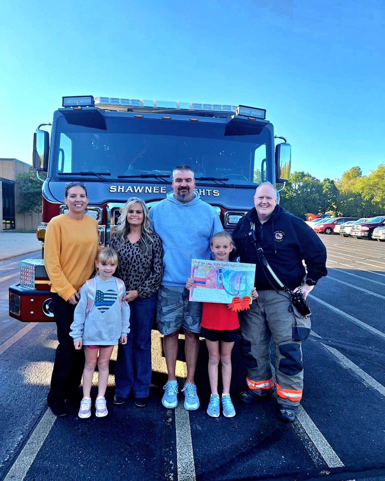Five adults and two children stand smiling in front of a fire truck labeled "Shawnee Heights." One child holds a colorful drawing, and one adult is in firefighter gear. The photo is taken outdoors in a parking lot.
