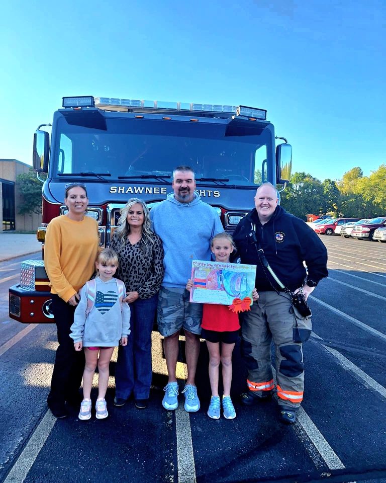Five adults and two children stand smiling in front of a fire truck labeled "Shawnee Heights." One child holds a colorful drawing, and one adult is in firefighter gear. The photo is taken outdoors in a parking lot.
