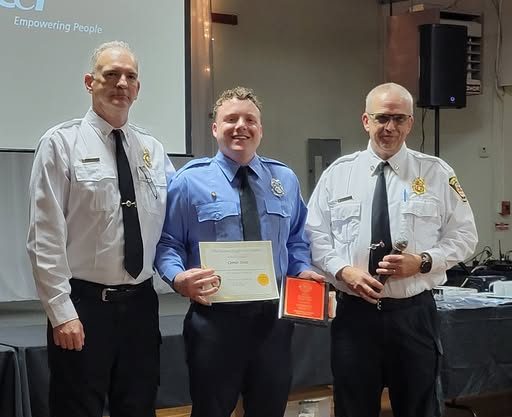 Three men in uniform stand indoors at an awards ceremony. The man in the center, in a blue shirt, holds a certificate and a red plaque. The two men in white shirts stand on either side, all smiling at the camera.