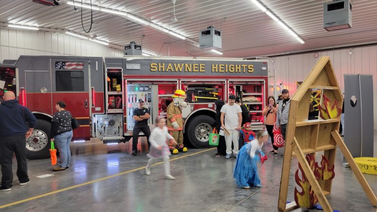 Children and adults enjoy activities inside a fire station near a Shawnee Heights fire truck. A person in a firefighter costume interacts with kids, and a wooden game structure with painted flames is set up in the foreground.
