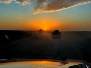View from inside a vehicle driving on a dusty road at sunset, with two trucks ahead and a dramatic, colorful sky with clouds illuminated by the sun.
