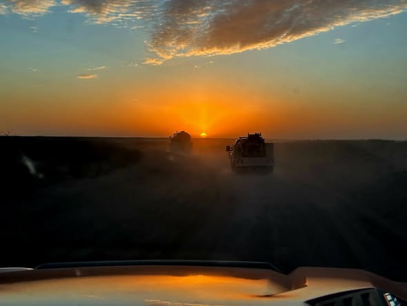 View from inside a vehicle driving on a dusty road at sunset, with two trucks ahead and a dramatic, colorful sky with clouds illuminated by the sun.