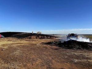 Firefighters in yellow gear stand on a scorched hill and beside a smoky area, working to contain a fire in a dry, open landscape under a clear blue sky.